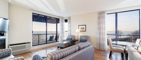 A modern living room with a sofa set, a dining table with chairs, a flat-screen TV, and large windows offering a view of the city.