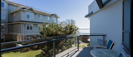 A balcony with a glass table and chairs, overlooking a residential area with houses and a lawn.