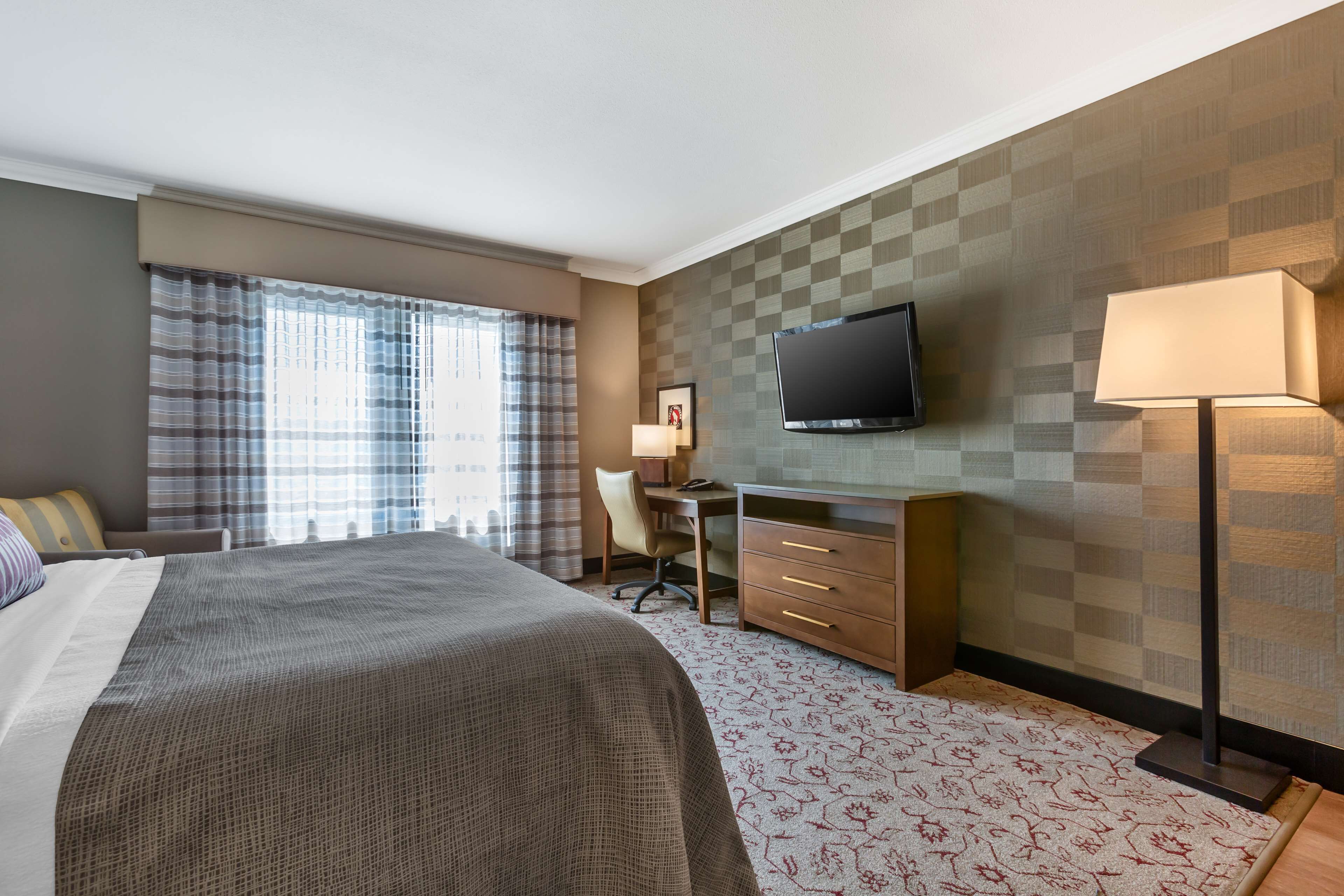 A hotel room with a large bed, a desk with a chair, a television mounted on the wall, and a lamp.