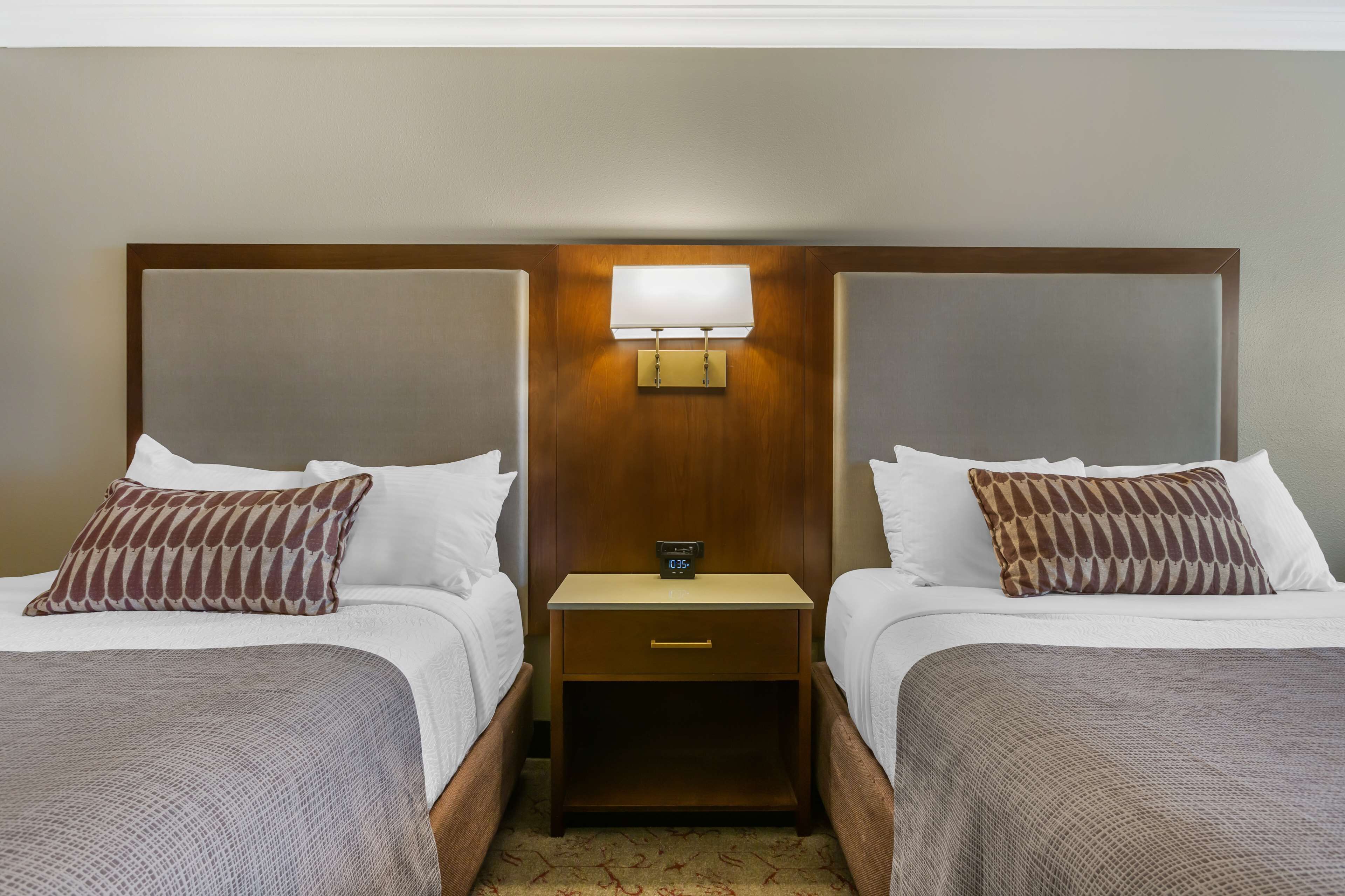 Une chambre d’hôtel avec deux lits, chacun agrémenté d’un oreiller à motifs, une table de chevet avec une horloge et un luminaire fixé au mur.