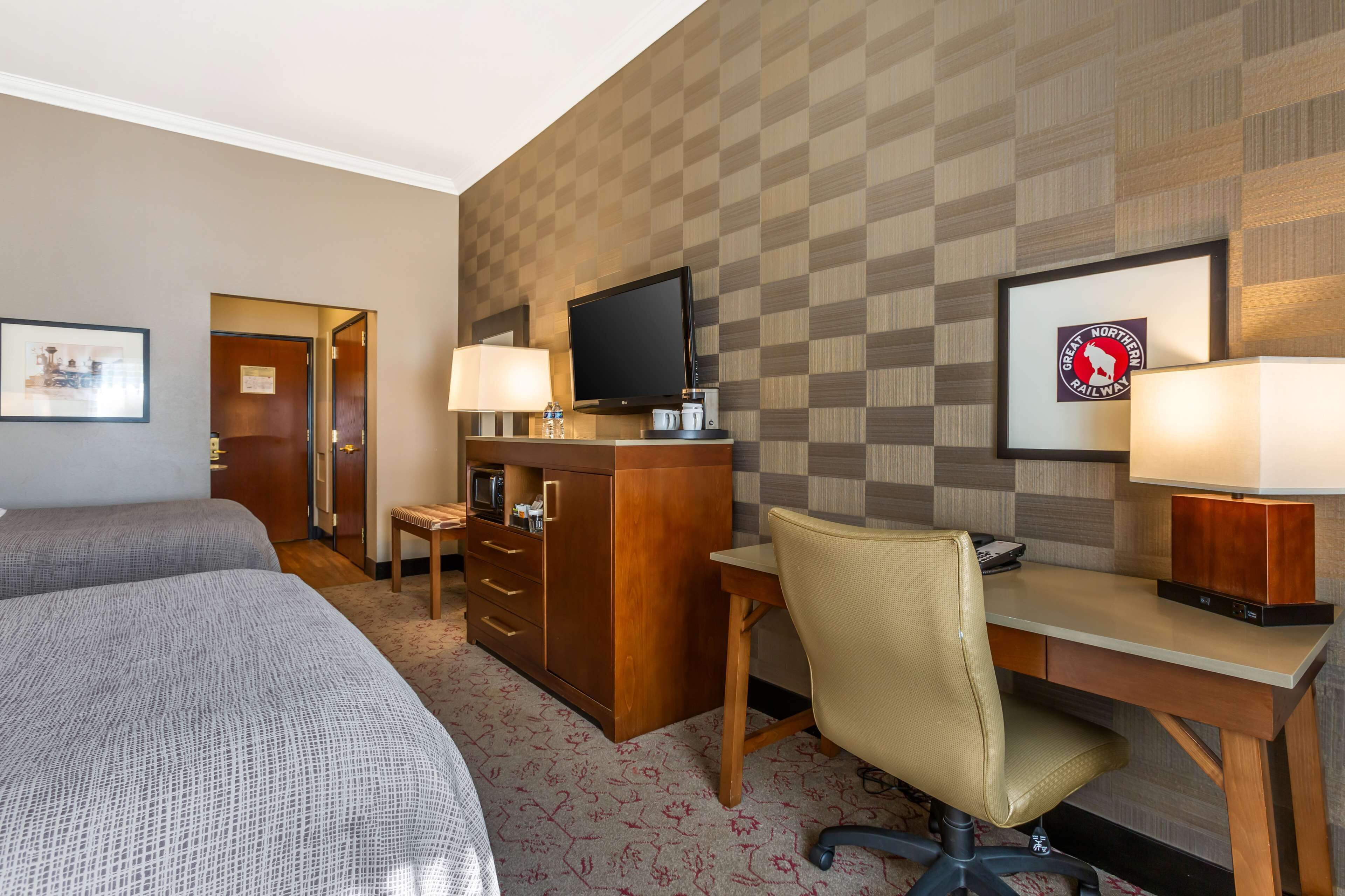 Ein Hotelzimmer mit zwei Betten, einem Schreibtisch mit Stuhl, einem Fernseher und einem gerahmten Logo an der Wand.
