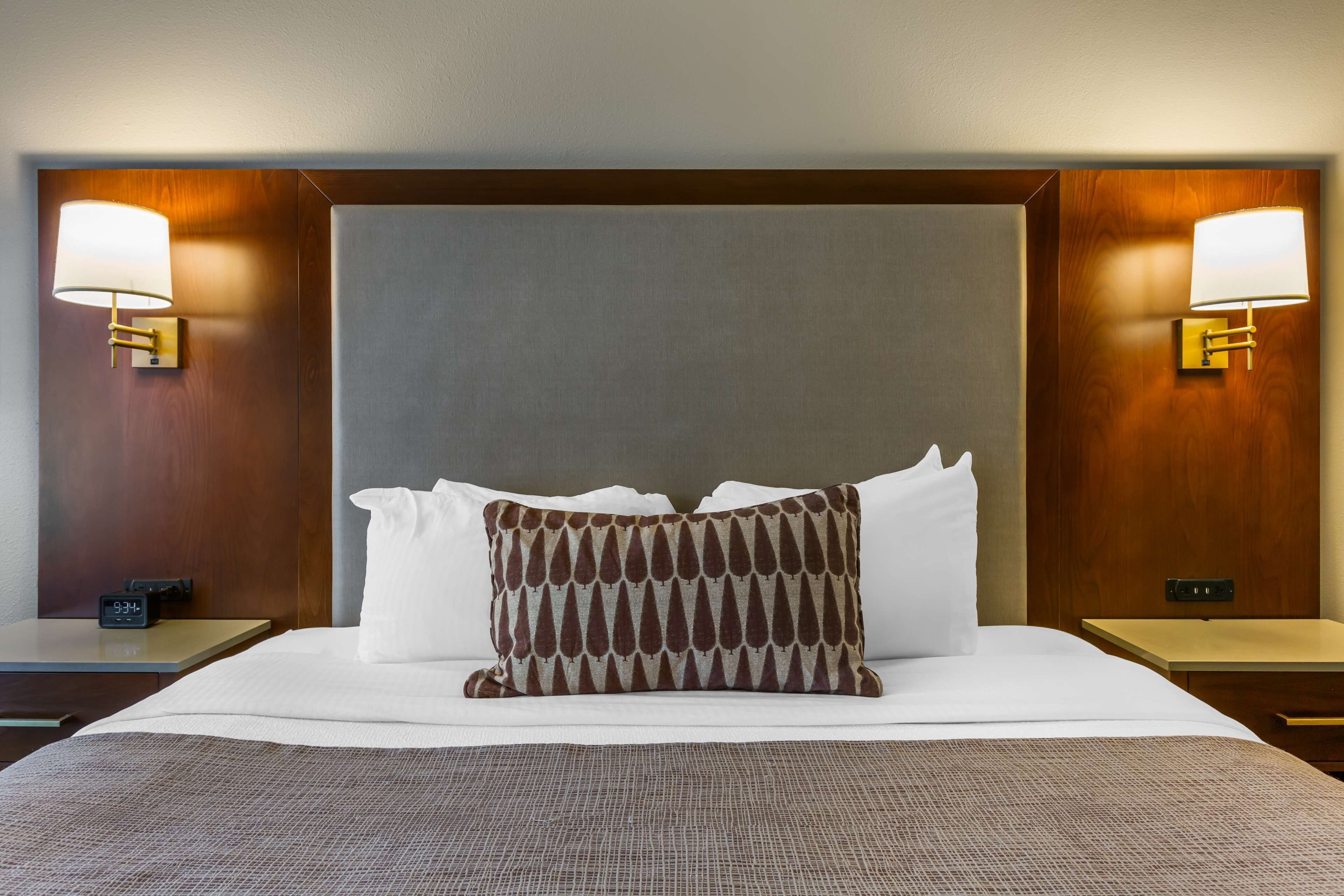 Habitación de hotel con una cama grande, dos mesitas de noche, una mesita auxiliar, un reloj de pared y dos lámparas de pared.