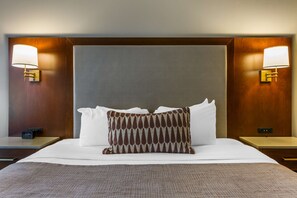 A hotel room with a large bed, two bedside tables, a nightstand, a wall clock, and two wall-mounted lamps.