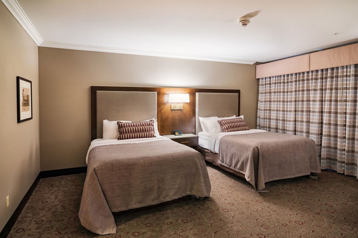 Een hotelkamer met twee bedden, een nachtkastje, een lamp, een lijst met een foto aan de muur en een raam met gordijnen.