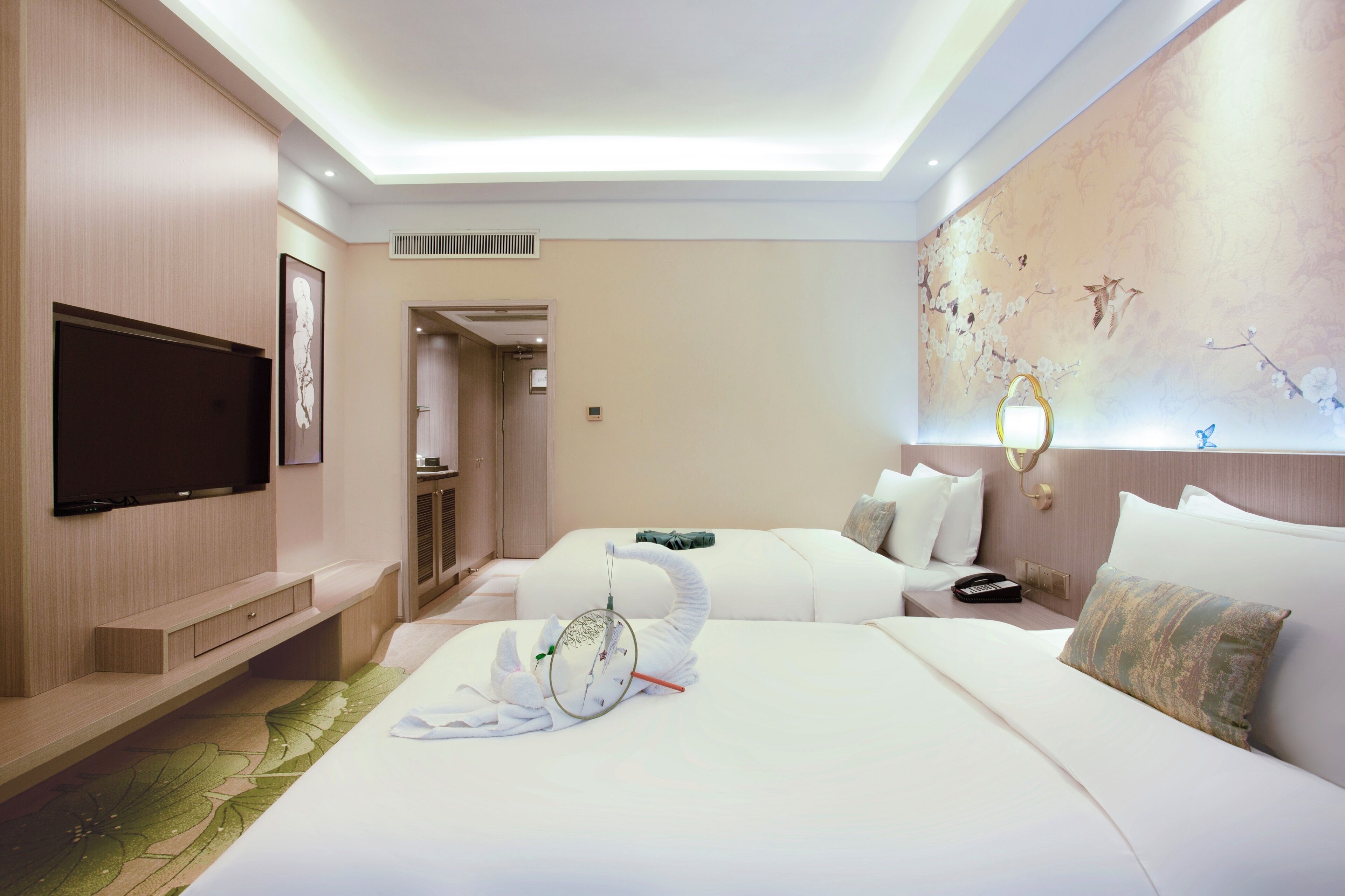 A hotel room with a flat-screen TV, two beds, a wall-mounted TV, a painting, and a bedside table with a lamp.