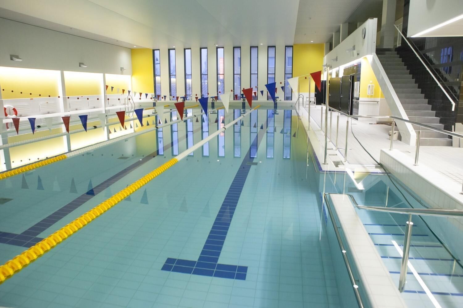 Una amplia piscina interior con varias pistas, una escalera a la derecha y una pared amarilla con ventanas.
