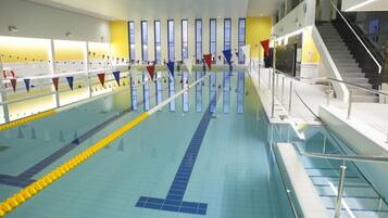 Una amplia piscina interior con varias pistas, una escalera a la derecha y una pared amarilla con ventanas.