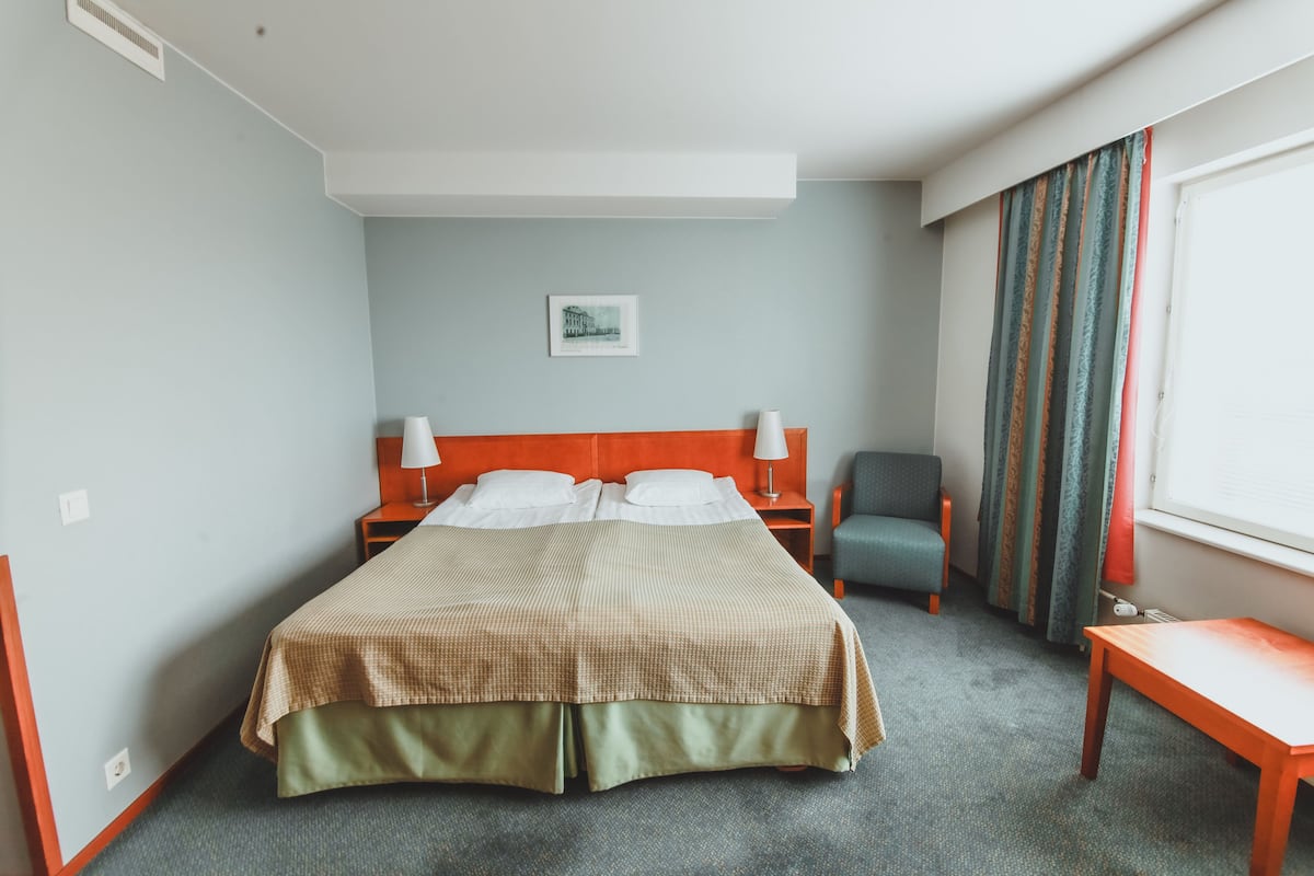 Una habitación de hotel con cama, dos mesitas de noche, una silla, un banco, una ventana con cortinas y un cuadro colgado en la pared.
