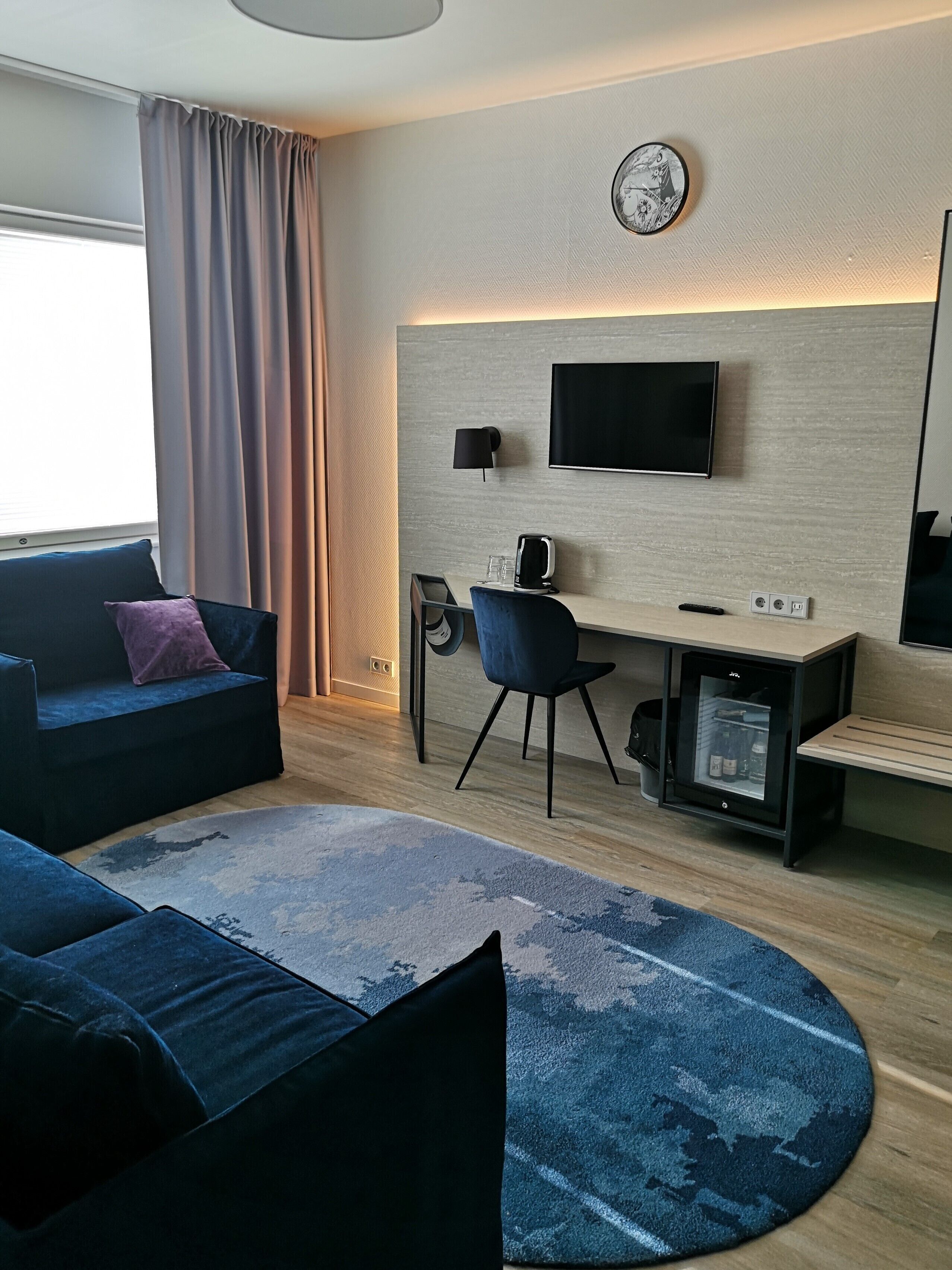 A modern hotel room with a sofa, a desk with a computer, a television, and a minibar.