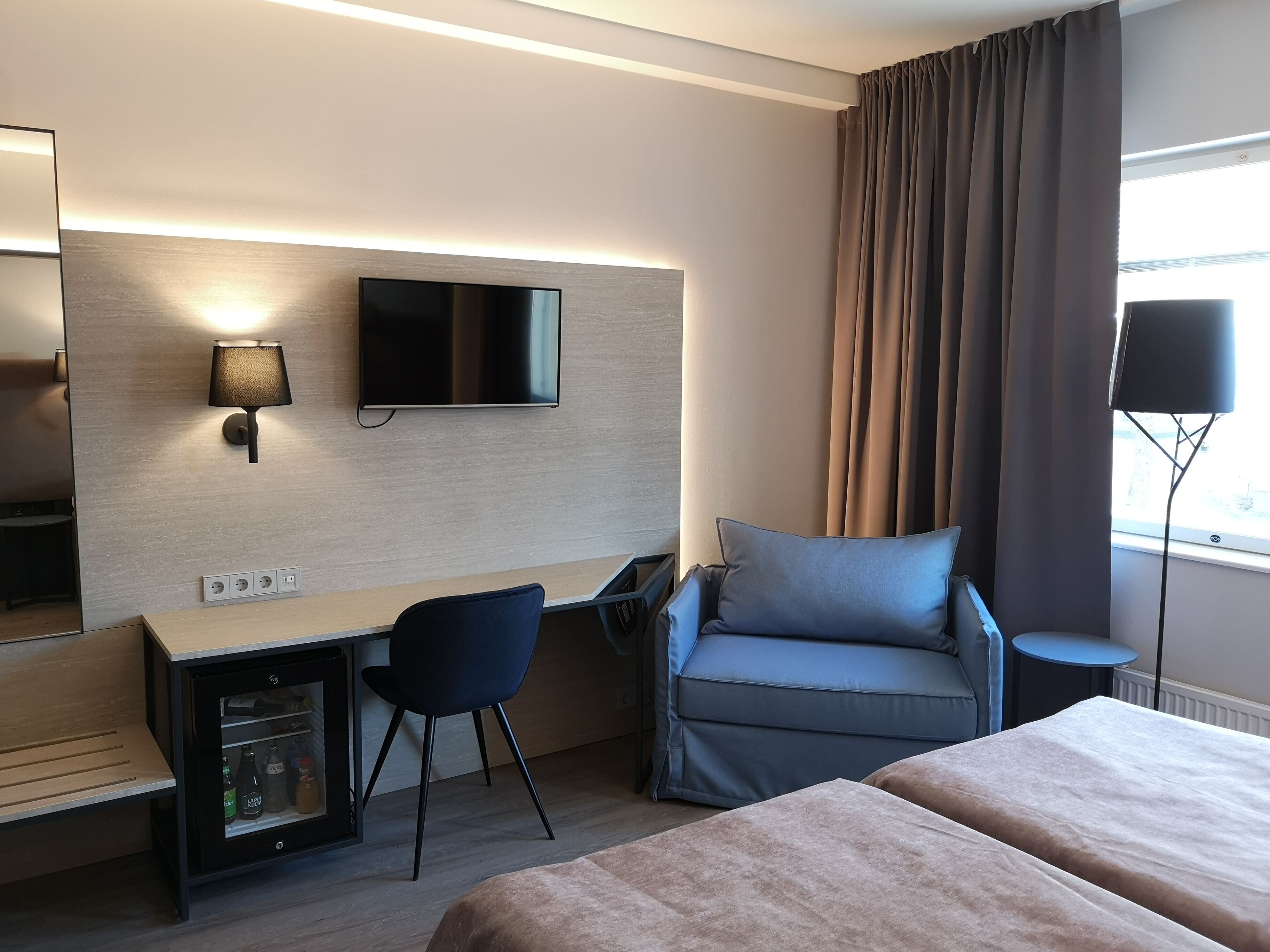 Une chambre d’hôtel comprenant un lit, un bureau avec une chaise, un téléviseur fixé au mur, une lampe et une fenêtre avec des rideaux.