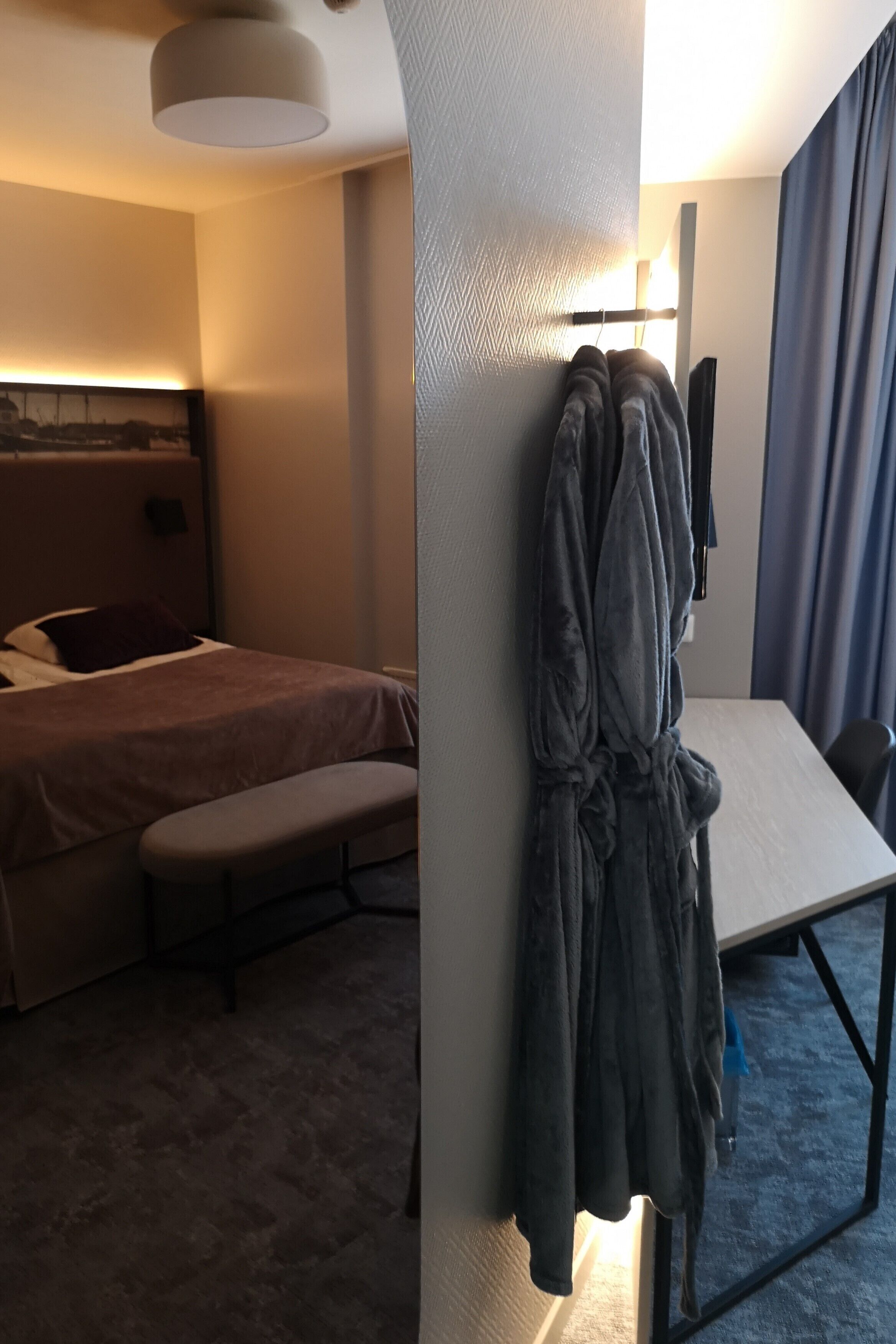 Habitación de hotel con cama, un banco, una bata colgada en la pared y un escritorio con silla.