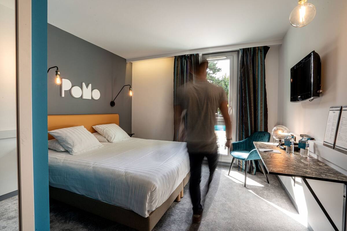 Une chambre d’hôtel avec un lit, un bureau et une chaise, une télévision et une fenêtre avec des rideaux.