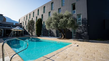 A swimming pool with a metal ladder, surrounded by a brick patio and a building with multiple windows and a tree.