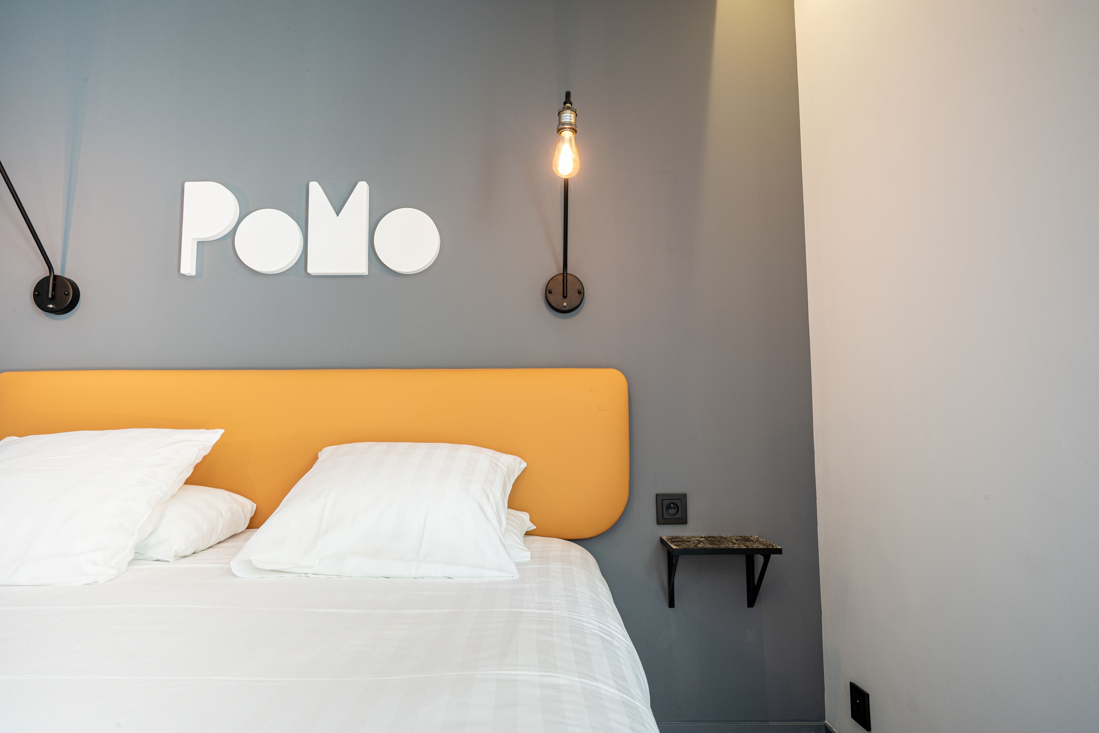 Un dormitorio con cama, almohadas blancas, una lámpara de pared y un estante pequeño.