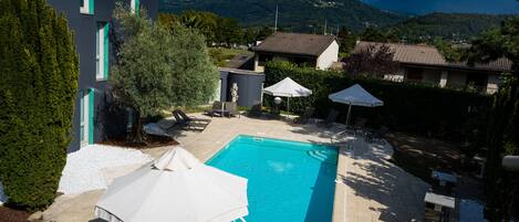 Ein Schwimmbecken mit Liegestühlen und Sonnenschirmen, umgeben von Gebäuden und Bergen.