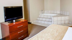 A hotel room with a flat-screen TV on a wooden dresser, a large bed with a patterned bedspread, and a corner bathtub with steps leading up to it. - Comfort Suites Gadsden Attalla (Attalla)