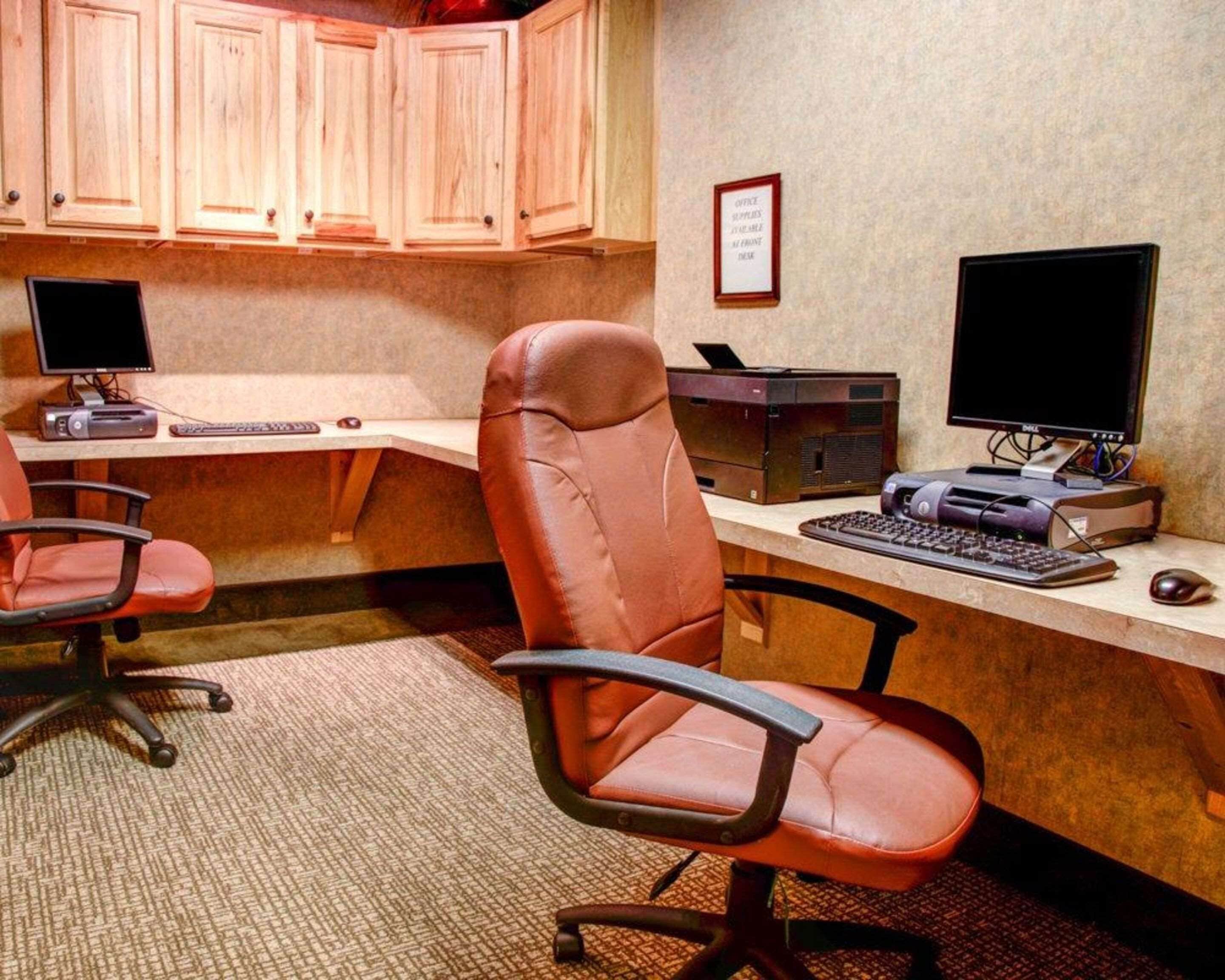 Un bureau à domicile équipé de deux bureaux, chacun comprenant un écran d’ordinateur, un clavier et une imprimante. Il y a deux chaises de bureau, une devant chaque bureau. La pièce est dotée de meubles de rangement en bois au-dessus des bureaux et d’un certificat encadré accroché au mur.