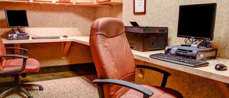 A home office with two desks, each with a computer monitor, keyboard, and printer. There are two office chairs, one in front of each desk. The room has wooden cabinets above the desks and a framed certificate on the wall.