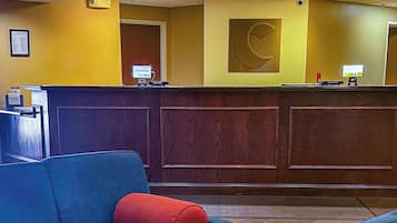 A reception area with a wooden reception desk, a blue chair with a red cushion, and a wall-mounted clock.