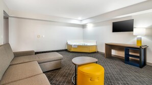 Una habitación de hotel moderna con sofá, una mesita pequeña, un otoman amarillo, un escritorio de madera, un televisor de pantalla plana y una bañera.