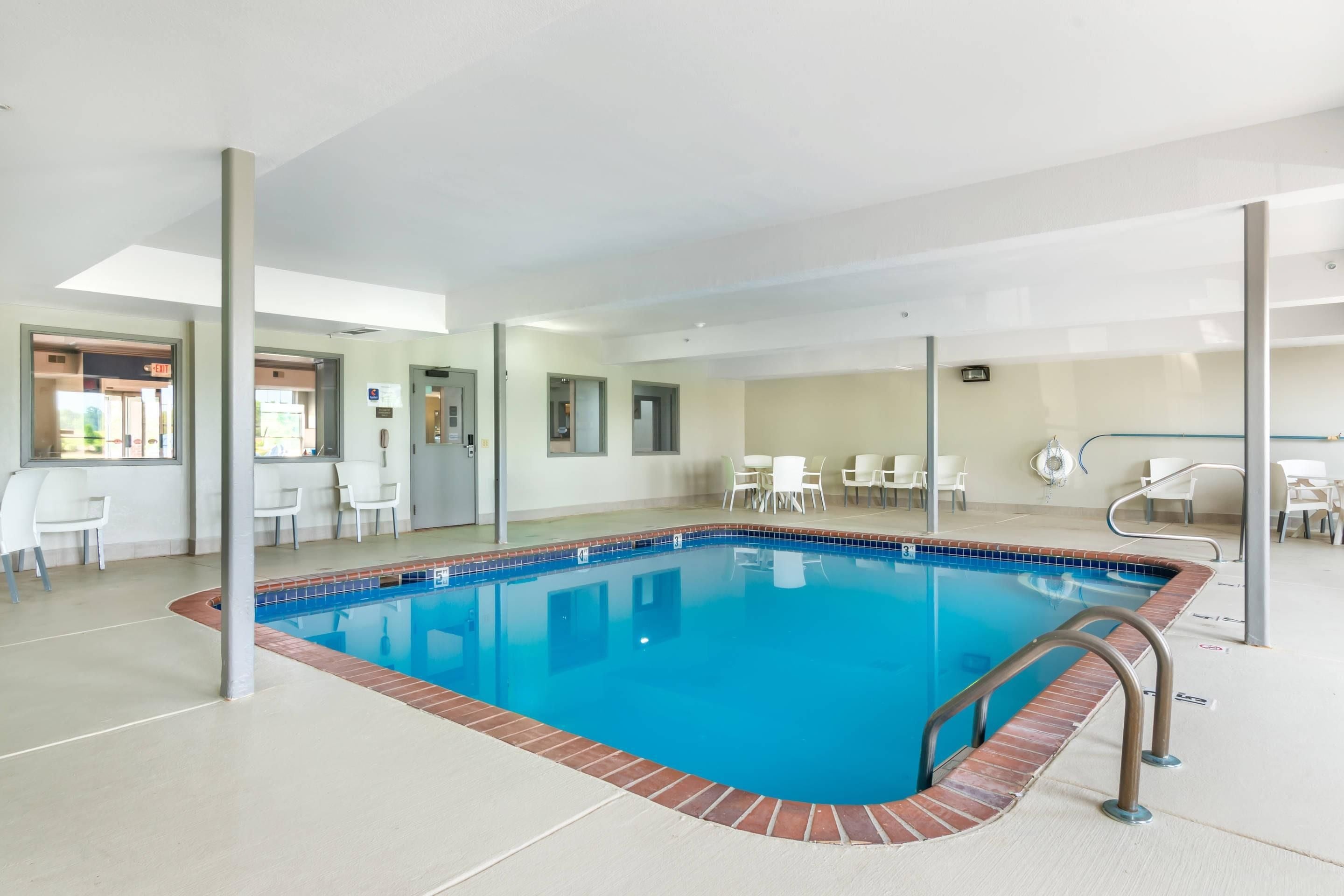 Una piscina interior con agua azul cristalina, rodeada de sillas y mesas blancas y un suelo de baldosas.