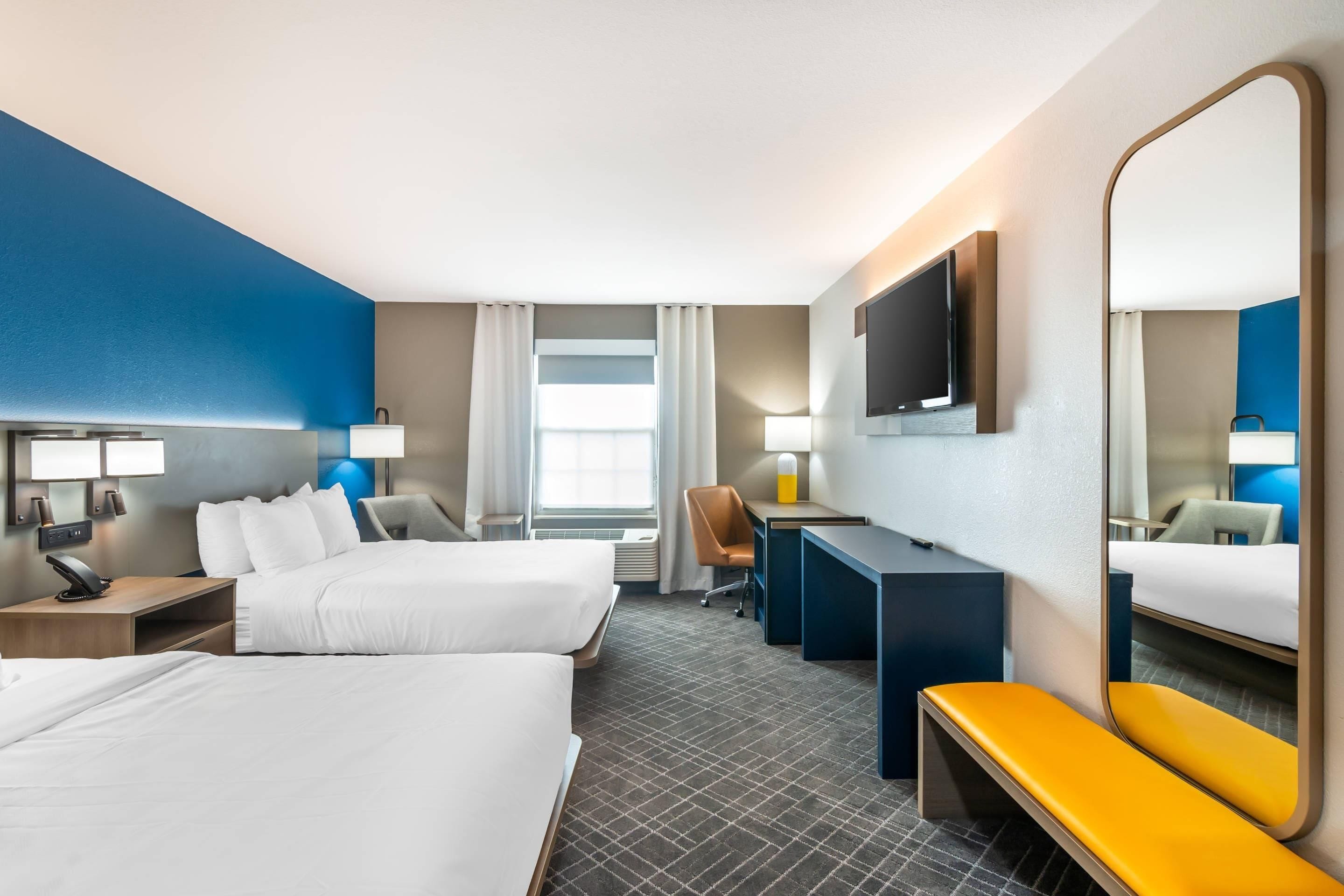 Habitación de hotel con dos camas, un escritorio, una silla, un banco, un televisor y un espejo grande.