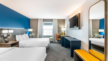 Habitación de hotel con dos camas, un escritorio, una silla, un banco, un televisor y un espejo grande.