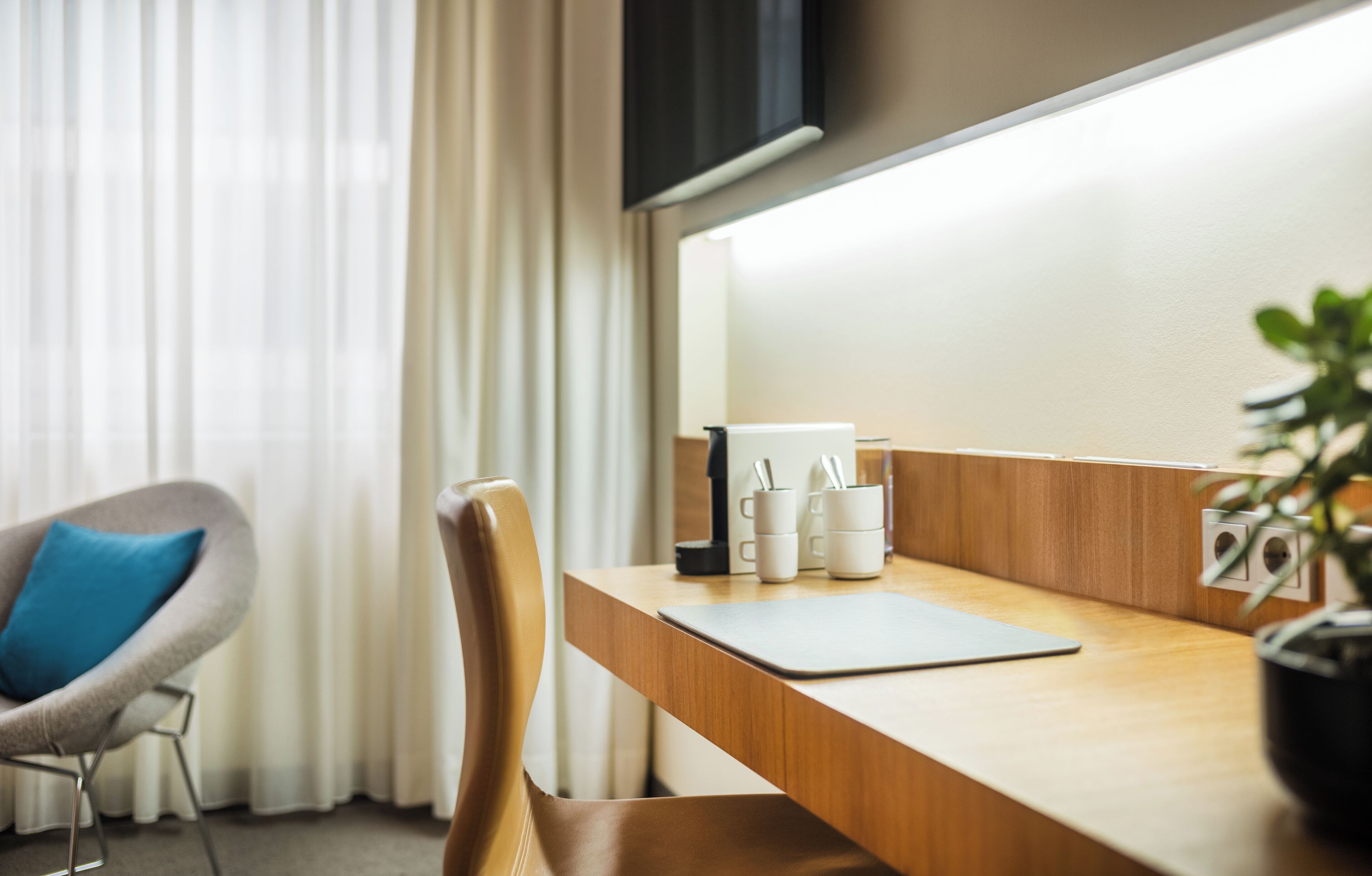 A hotel room with a wooden desk, a chair, a potted plant, and a window with curtains.