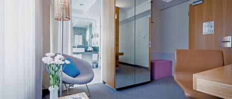A modern hotel room with a wooden desk, a comfortable armchair, a large mirror, and a vase with flowers.