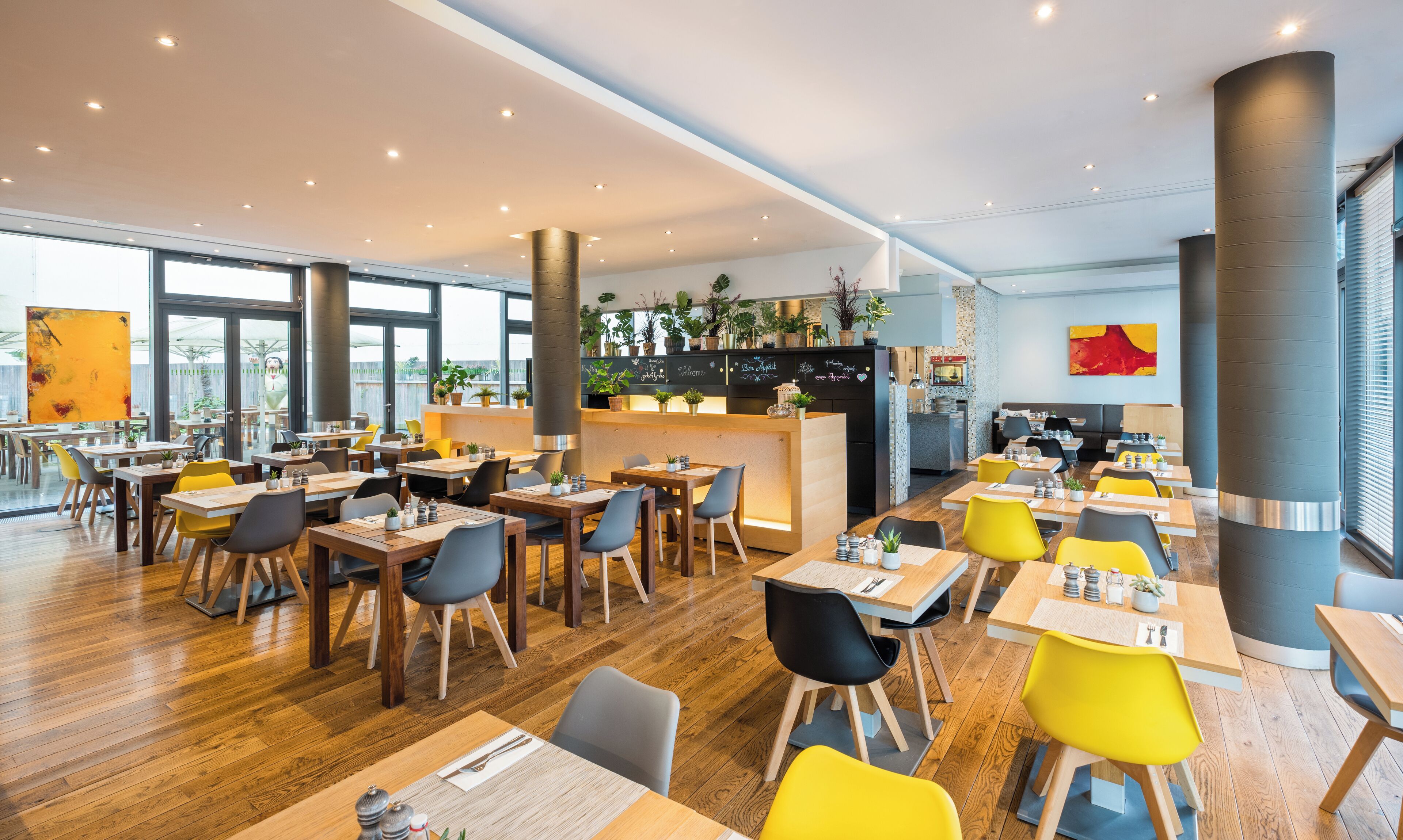A modern restaurant with wooden tables and chairs, a central bar area with plants, and large windows allowing natural light.