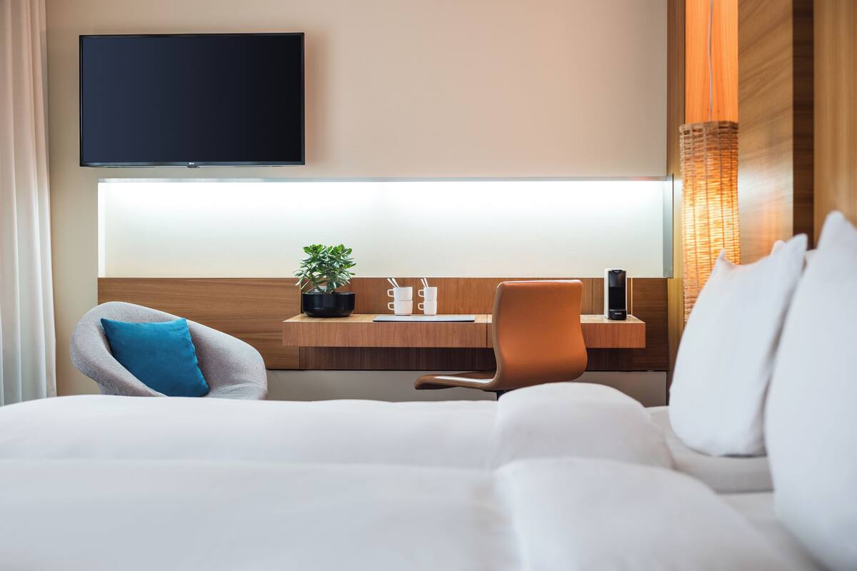 A hotel room with a bed, a desk with a plant, a chair, a TV, and a bedside table with a lamp.
