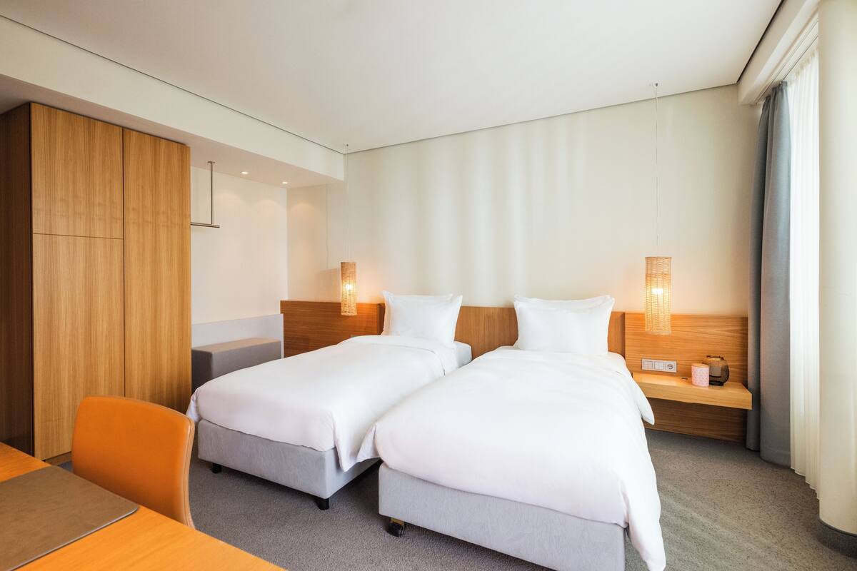A hotel room with two beds, a wooden wardrobe, a desk with a chair, and a bedside table with a lamp.