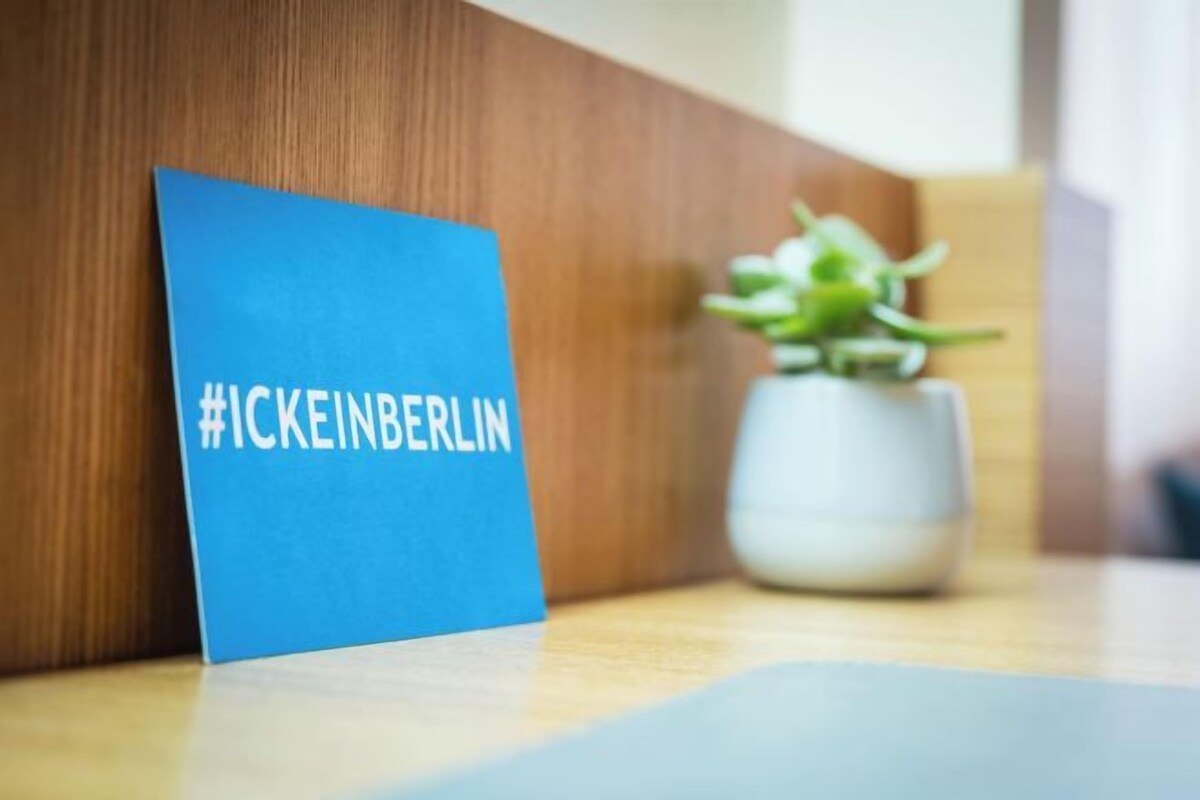 A blue card with the text "#ICKEINBERLIN" on a wooden surface next to a white mug with a green plant.