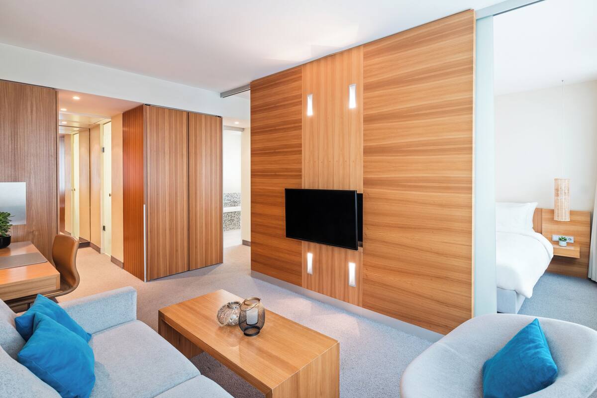A modern hotel room with a wooden accent wall, a flat-screen TV, a sofa, a coffee table, and a bed with a nightstand.