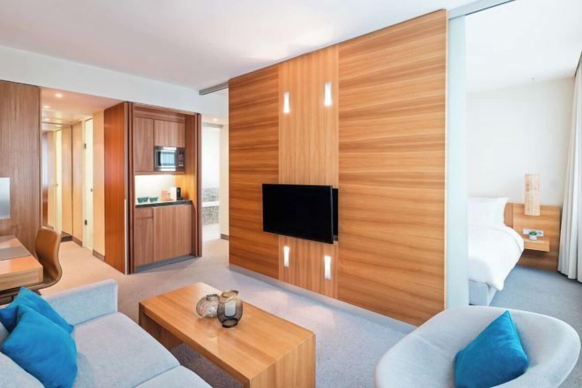 A modern hotel room with a wooden accent wall, a flat-screen TV, a sofa, a coffee table, and a bed with a nightstand.