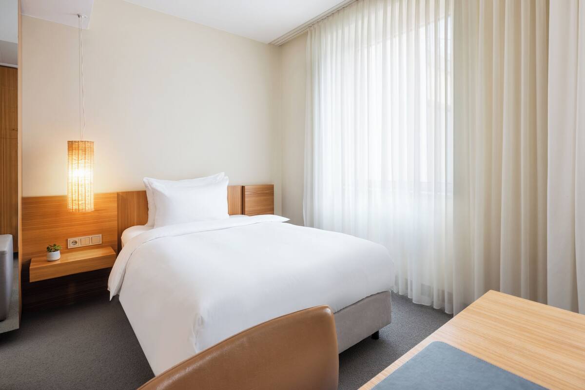 A hotel room with a bed, a bedside table with a lamp, a chair, and a view of a window with sheer curtains.