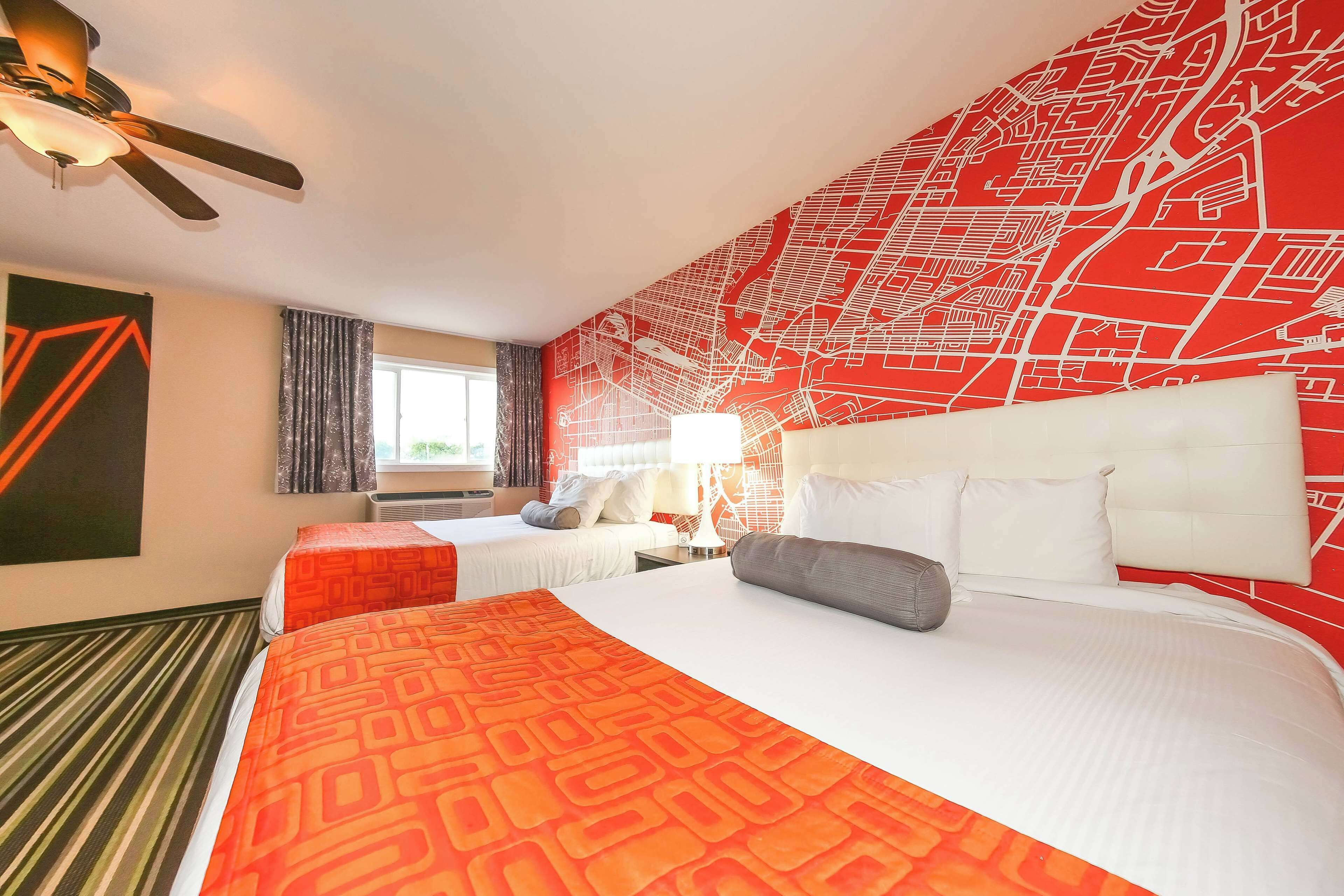 A hotel room with two beds, a large bed with an orange patterned bedspread and a smaller bed with a white bedspread. There is a red and white wall mural, a ceiling fan, and a window with curtains.