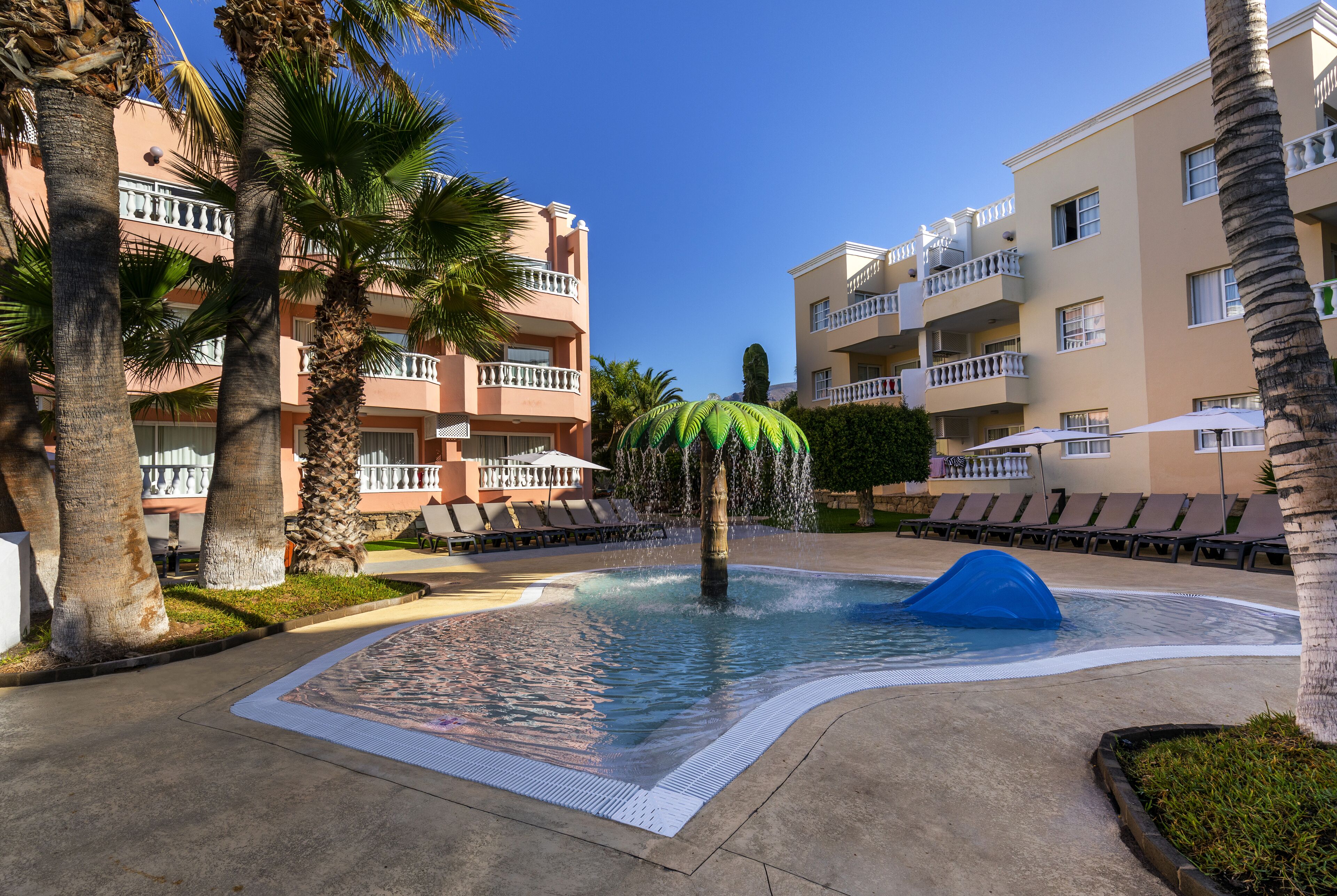 Une piscine avec un toboggan bleu, entourée de palmiers et de chaises longues, avec des immeubles à plusieurs étages en arrière-plan.