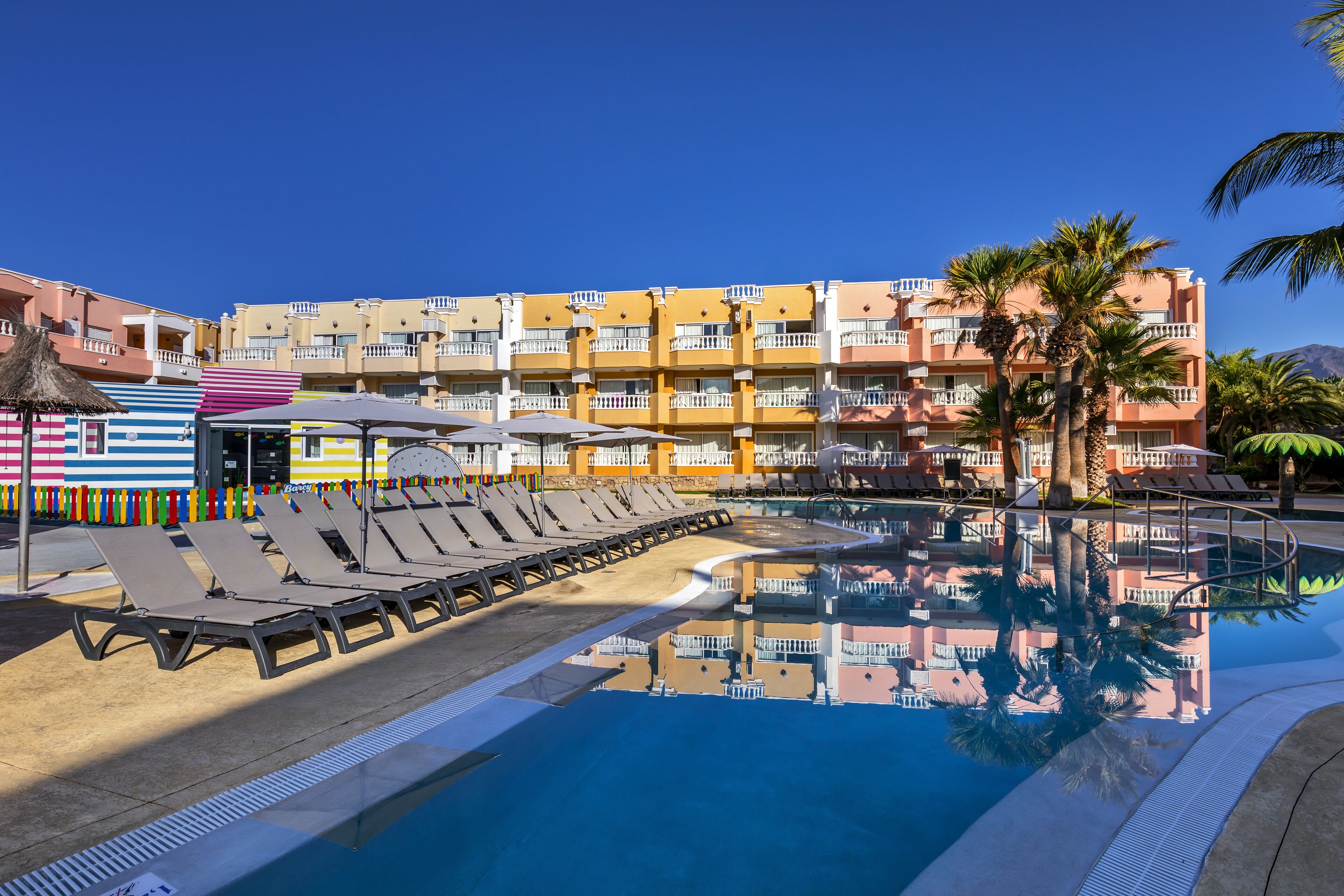 Un área de piscina hotelera con sillones de descanso y sombrillas, rodeada de edificios coloridos y palmeras.