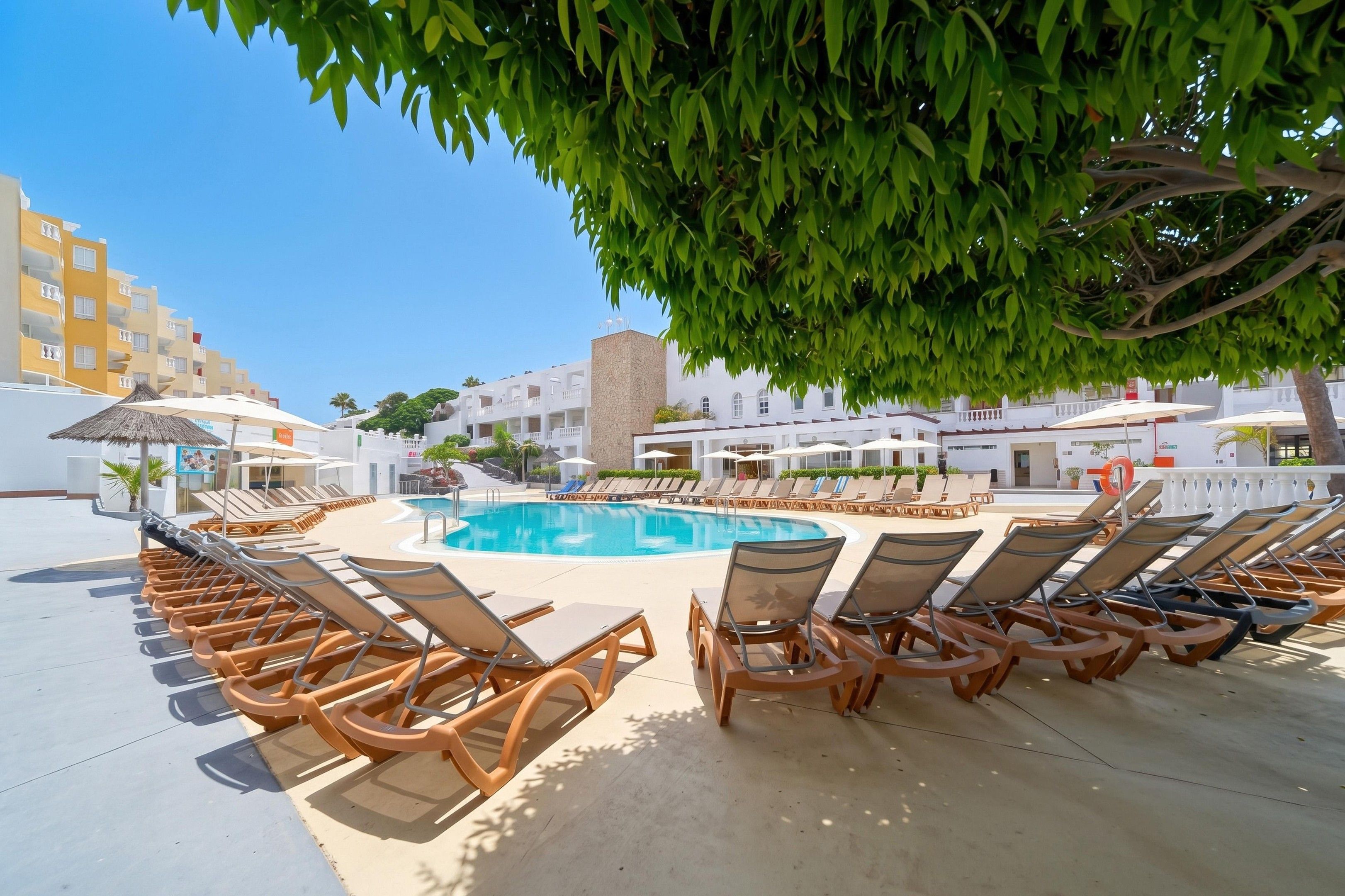 A poolside area with sun loungers and umbrellas, surrounded by buildings with balconies.