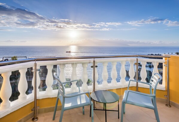 Ein Balkon mit zwei Stühlen und einem kleinen Tisch, der Blick auf die Küste mit Meer und einem Boot bietet.