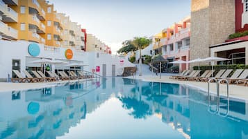 Una piscina con tumbonas y sombrillas, rodeada de edificios de varios pisos con balcones.