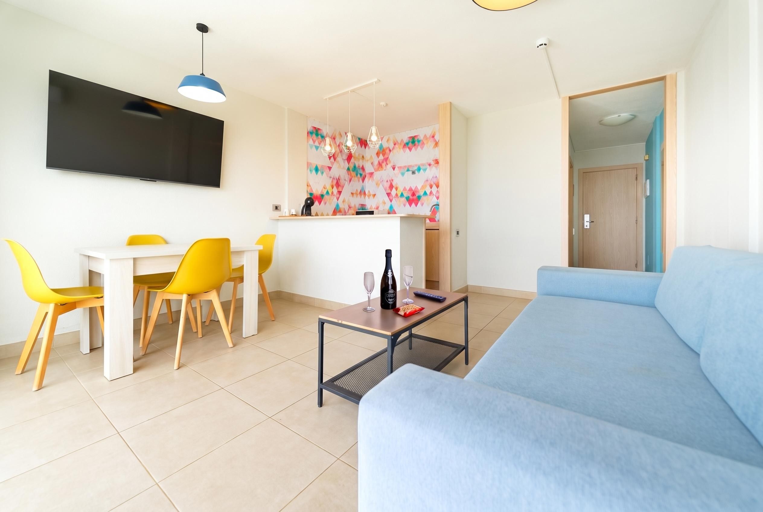 A modern living room with a blue sofa, yellow chairs, a dining table, and a TV mounted on the wall.