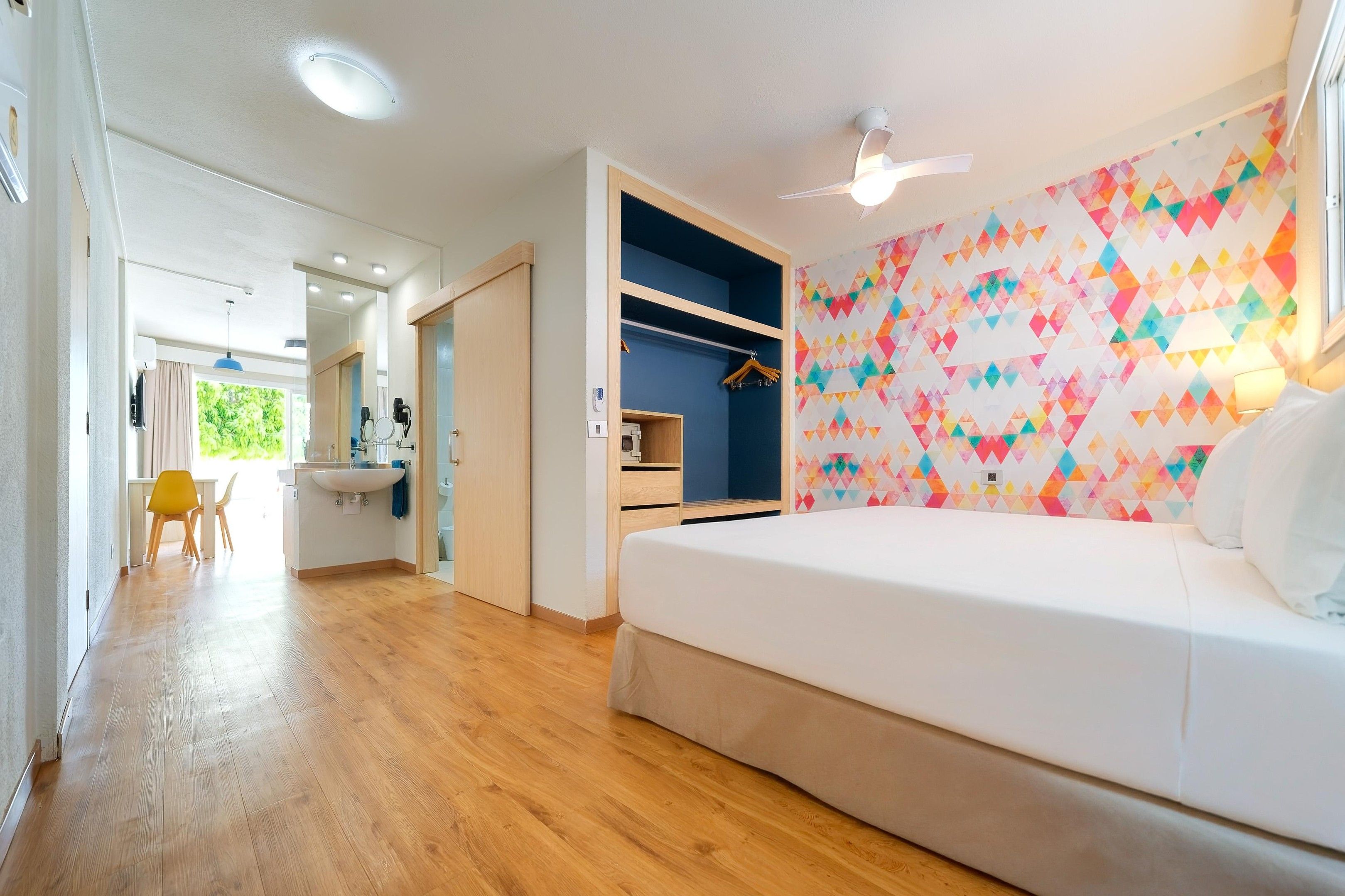 Een moderne slaapkamer met een bed, een plafondventilator, een badkamer met een wastafel en een kleurrijk geometrisch wandontwerp.