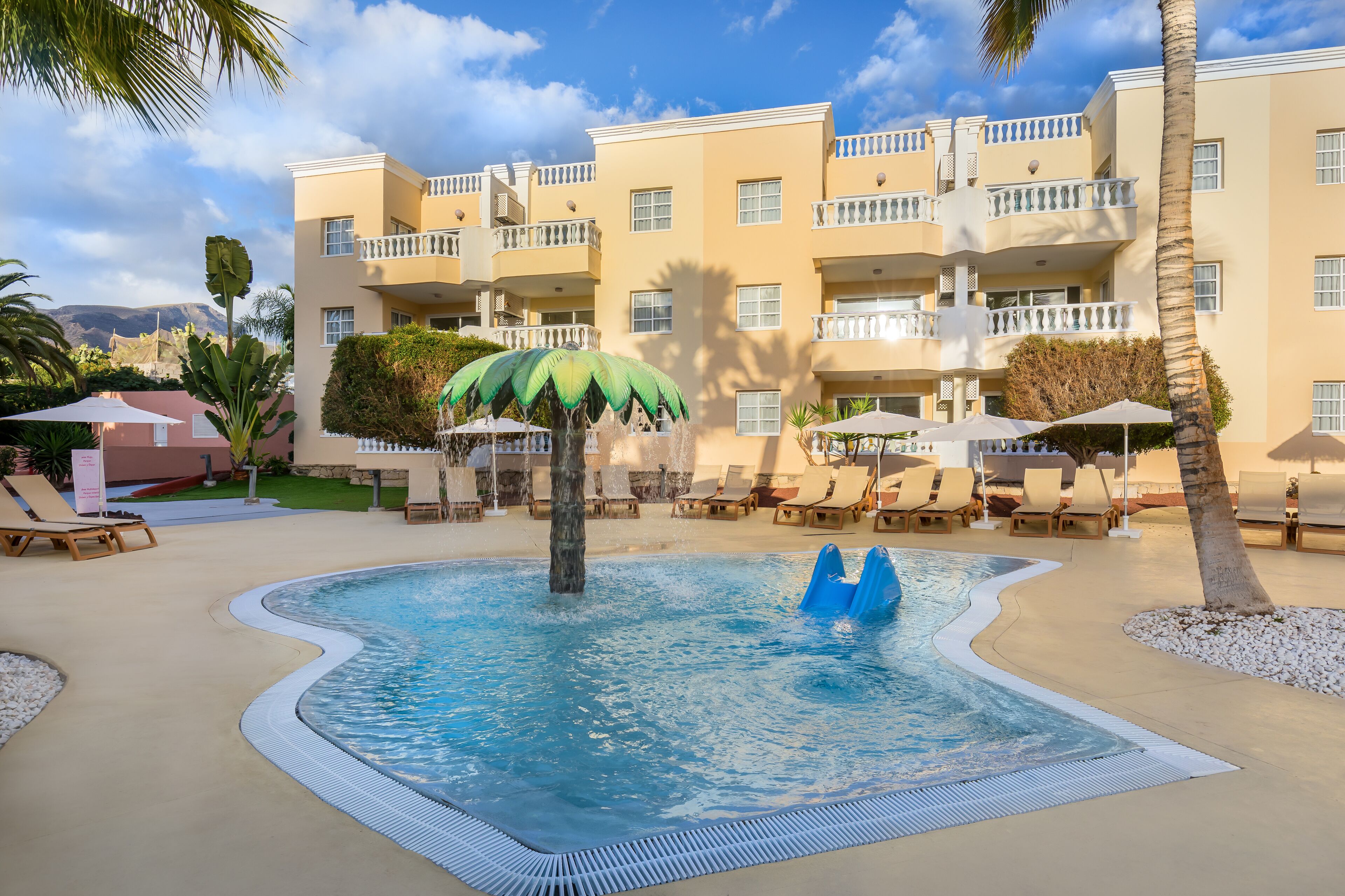 Un espace piscine doté de chaises longues et de parasols, entouré d’un bâtiment avec des balcons.