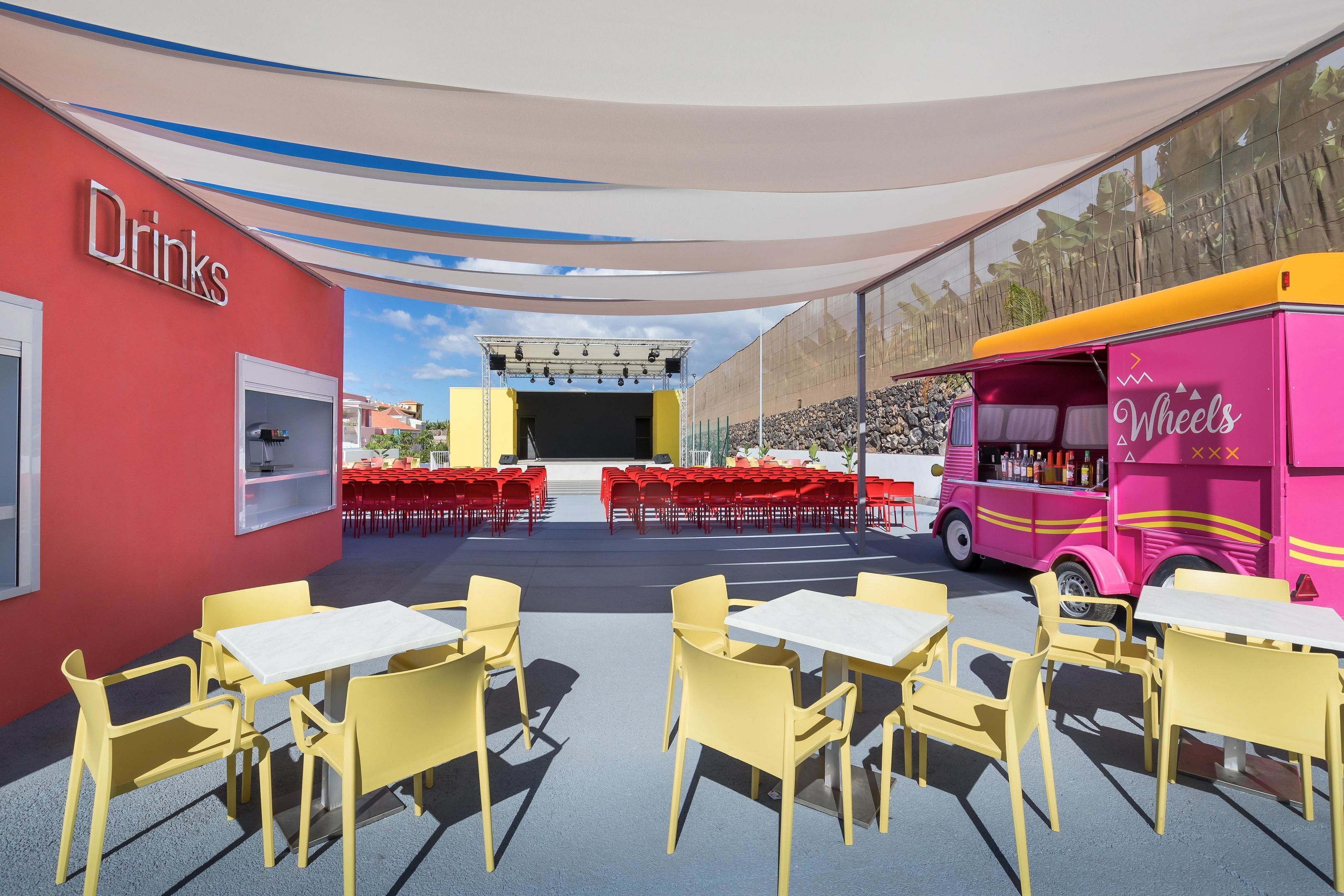 An outdoor dining area with yellow tables and red chairs, a pink food truck labeled 'Wheels,' and a stage with lighting equipment.