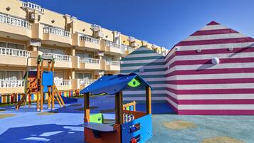 Un área de juegos infantiles con una estructura pequeña azul y naranja, un edificio a rayas y un hotel de varios pisos con balcones al fondo.