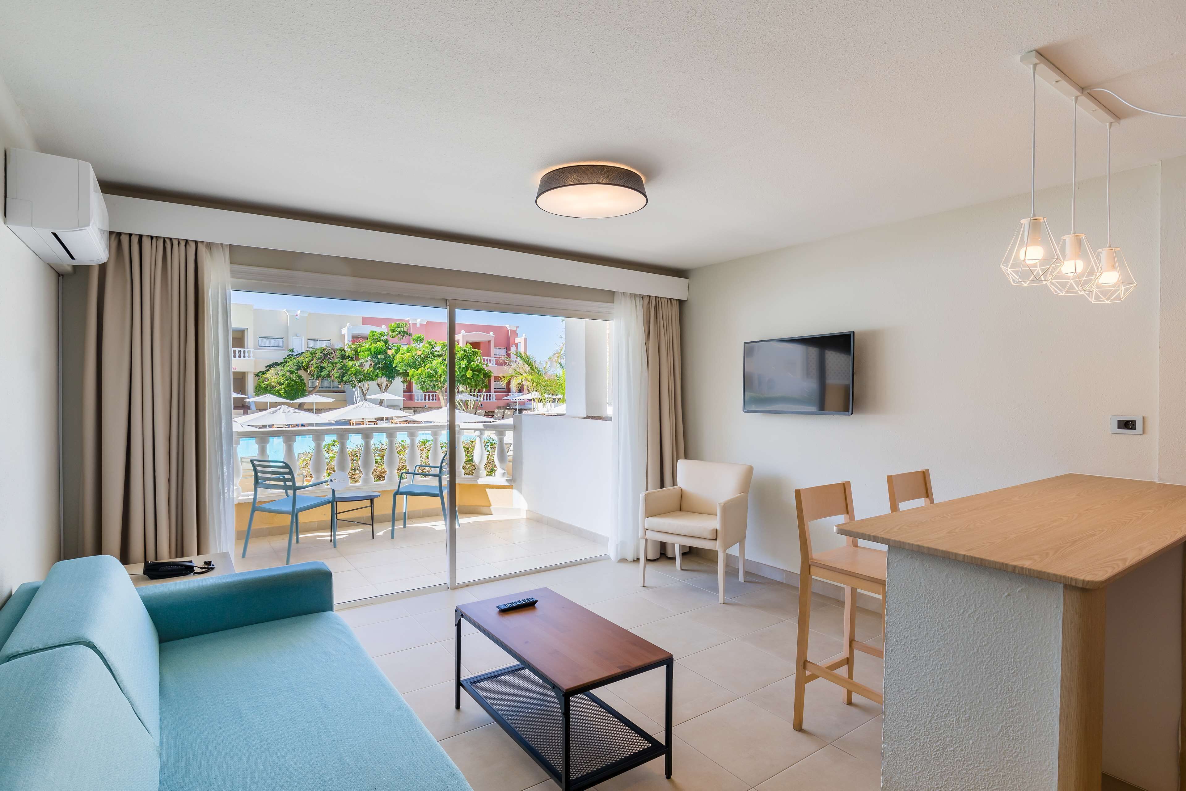 Een moderne hotelkamer met een bank, een kleine tafel, een eethoek met stoelen, een aan de muur bevestigde televisie en een balkon met uitzicht op gebouwen en bomen.