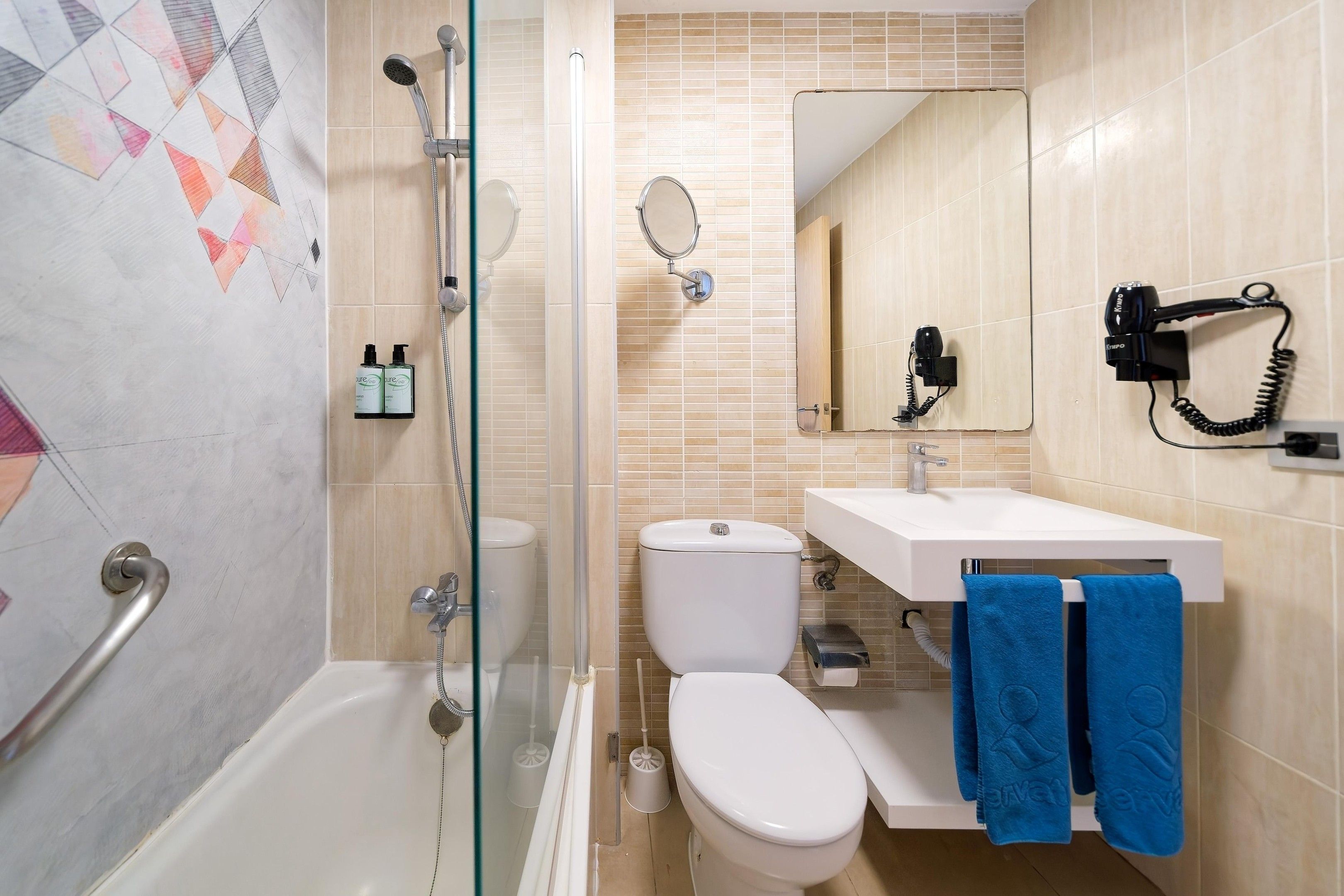 Un baño con ducha envuelta en vidrio, un inodoro, un lavabo con toallero y un secador de pelo montado en la pared.