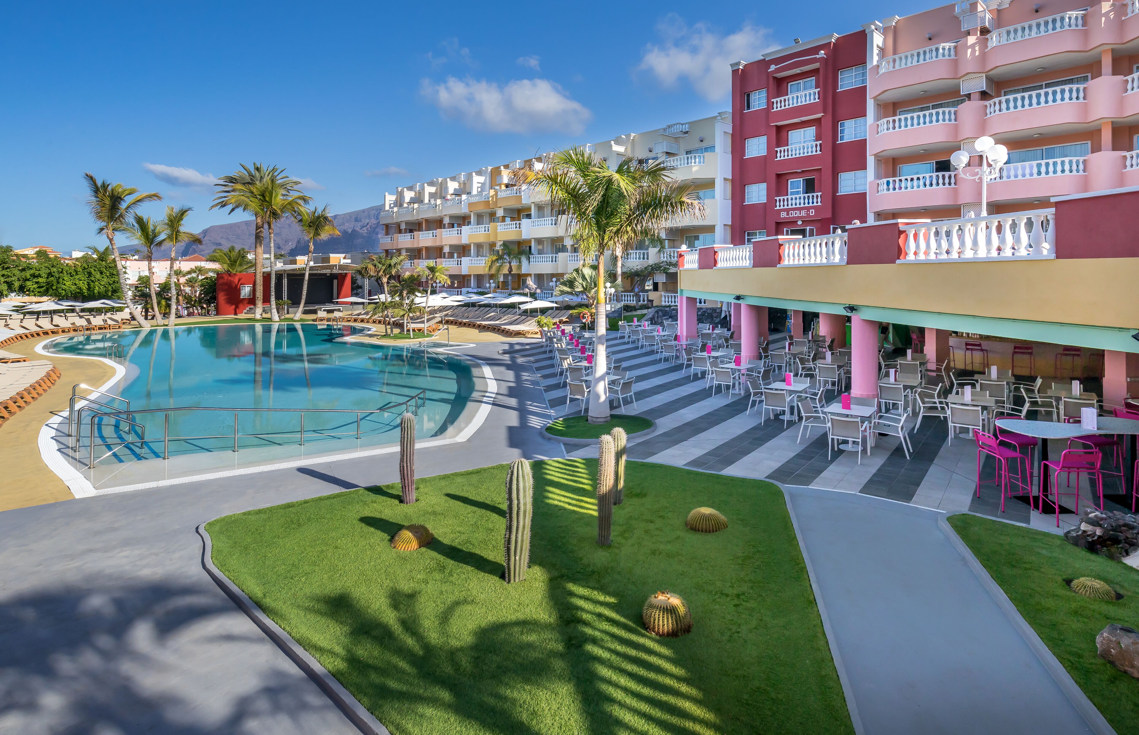 Zona de piscina de hotel con una piscina azul cristalina, palmeras y un edificio con balcones.